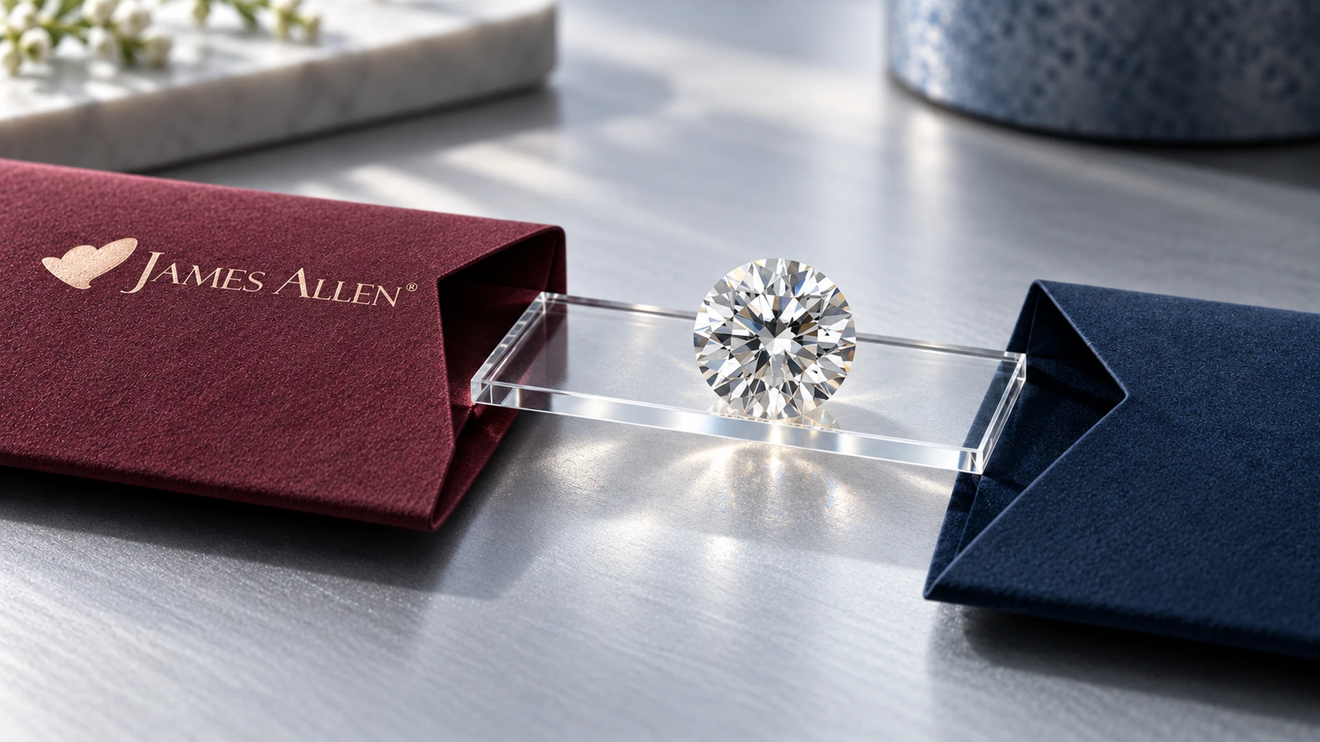 Round diamond on a clear acrylic bridge beside a burgundy box with the James Allen heart logo and a navy box